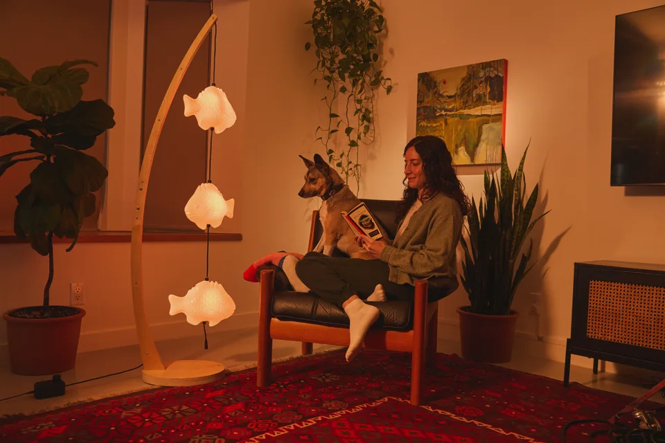 Three warm glowing fish on a curved wooden floor lamp beside a woman and her dog in a cozy living room